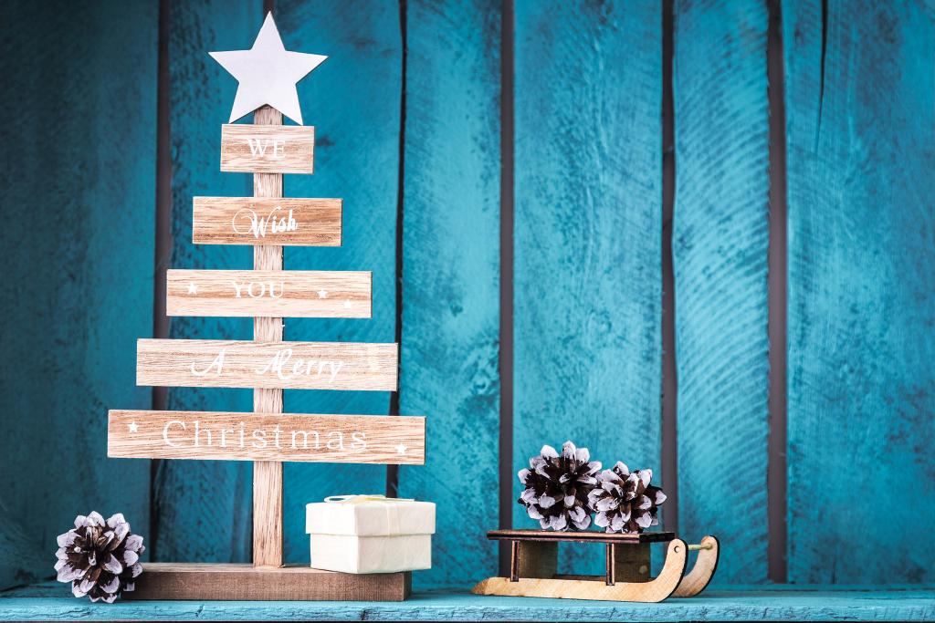 A wooden Christmas tree with a star topper and the words We wish you a Merry Christmas stands on a blue wooden surface beside a small white gift box, pinecones, and a toy sled, against a blue textured background.