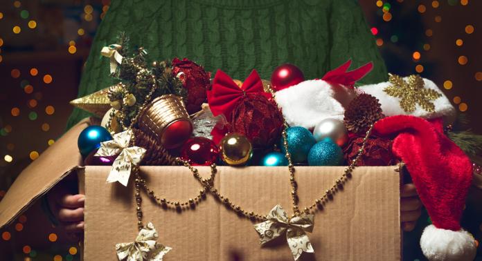 A person in a green sweater holds a cardboard box filled with Christmas ornaments, including baubles, pinecones, a Santa hat, bows, gold beads, and other festive decorations. Blurred holiday lights are in the background.