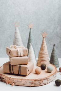 Three gifts wrapped in brown paper and string sit on a wooden tray, surrounded by decorative yarn trees with star toppers and wool balls, creating a cozy, minimalist holiday scene.