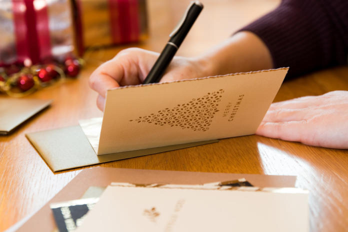 A person writes a message inside a Christmas card with a gold envelope on a wooden table, with wrapped gifts and festive decorations in the background.
