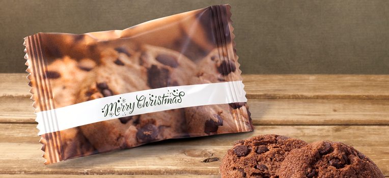 A cookie wrapped in festive packaging with Merry Christmas written on it sits on a wooden surface. Two chocolate chip cookies are placed nearby.