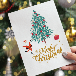 A hand holds a Christmas card with a drawing of Santa, a decorated tree, a red gift, and gold lettering that says “Merry Christmas.” A decorated Christmas tree is visible in the blurred background.