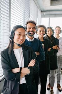 Five young professionals, some wearing headsets, stand in a line in a bright office with large windows. The woman in front smiles with her arms crossed; the group appears confident and ready to assist.